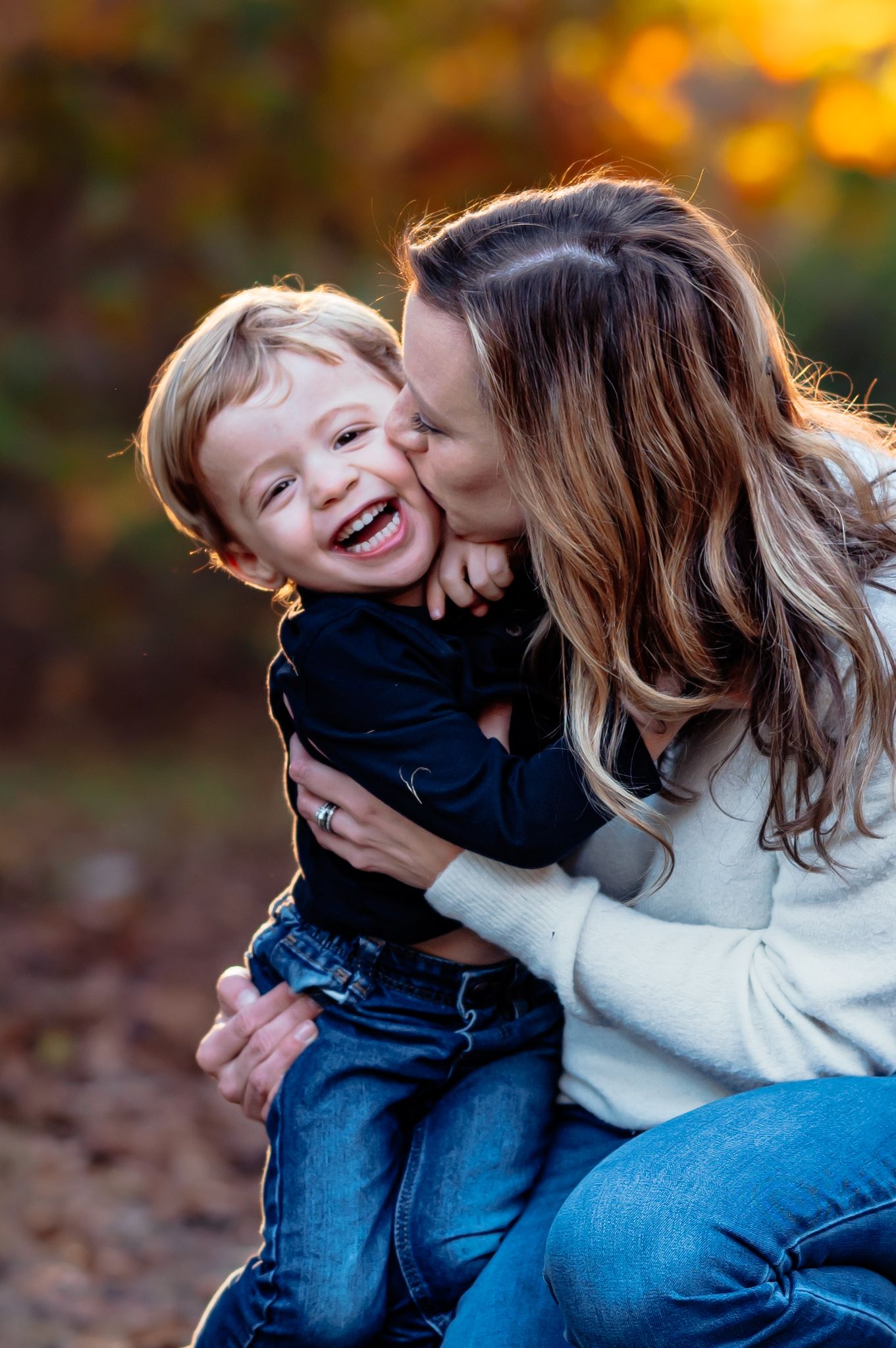 mother kneeling down to toddlers level to give a hug and kiss on the cheek to smiling boy during outdoor fall family photo session