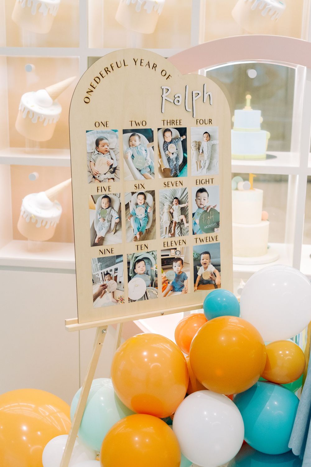 A pastel colored milestone photo display with monthly baby photos surrounded by orange, white and teal balloons.