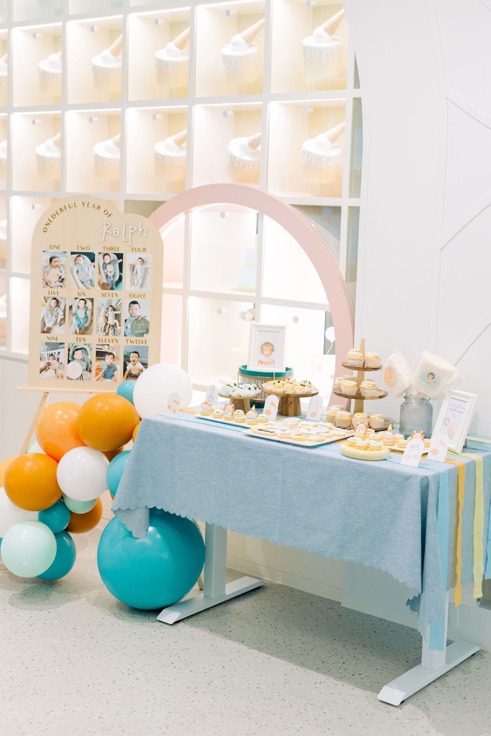 Modern dessert table setup with blue tablecloth, orange and teal balloons, and tiered cake stands at a party.