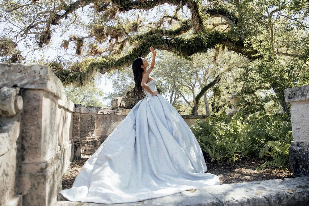 Couple in formal wedding attire pose together in a garden setting with stone architecture and natural lighting.