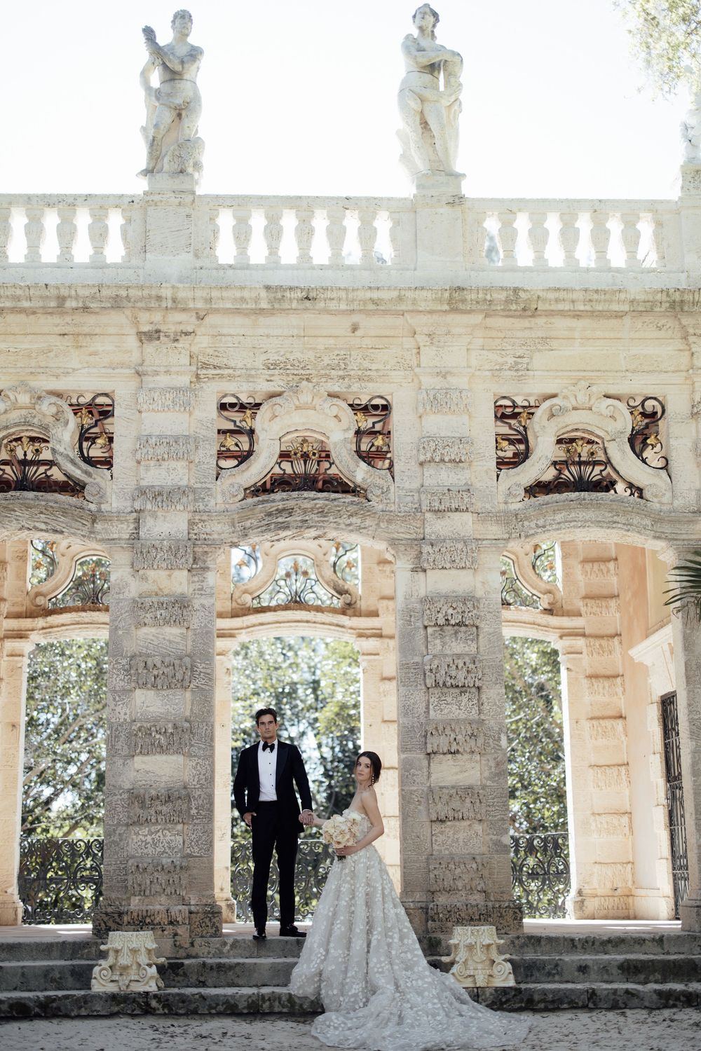 Couple in elegant wedding attire pose beneath ornate classical stone colonnade with decorative statues on top.