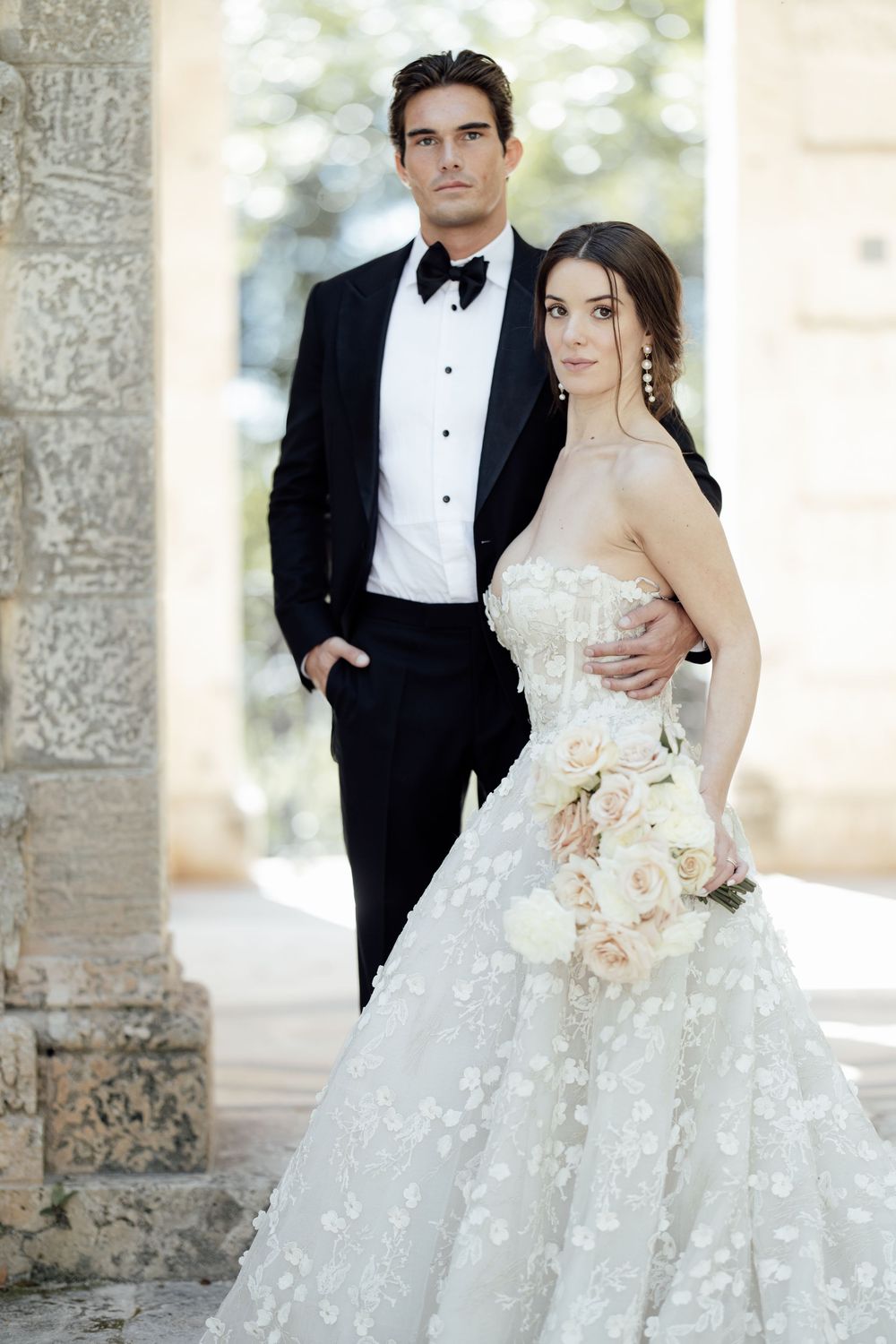Elegant wedding couple poses against stone columns in formal attire with peach bouquet.