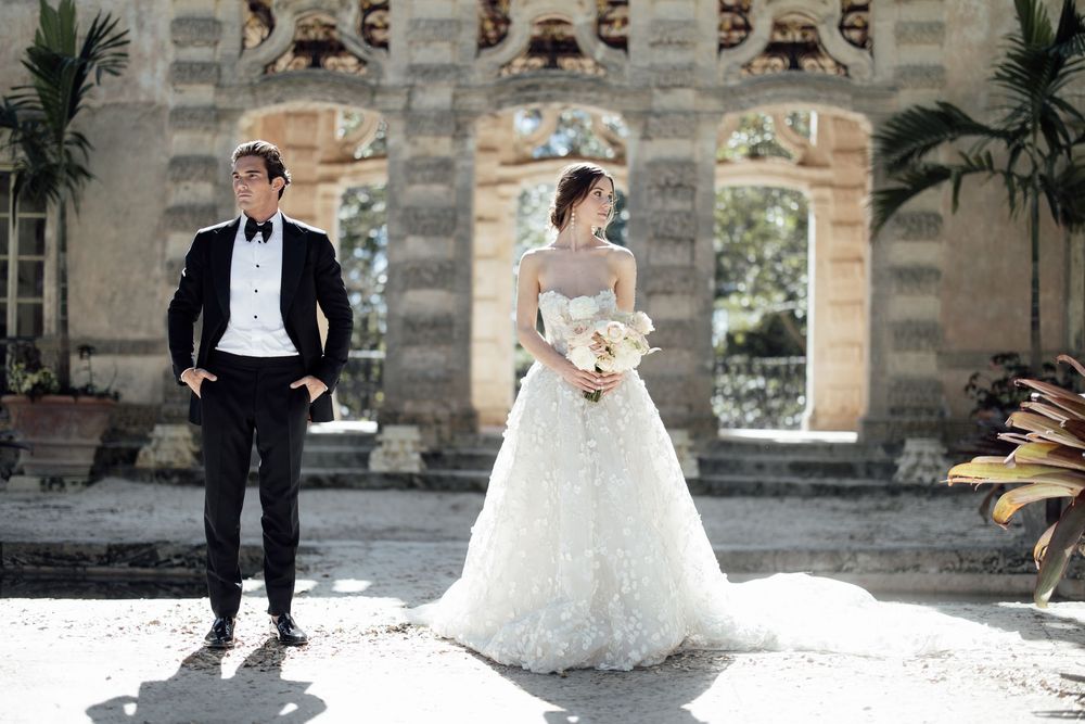 Wedding couple stands under outdoor stone archways surrounded by tropical plants.
