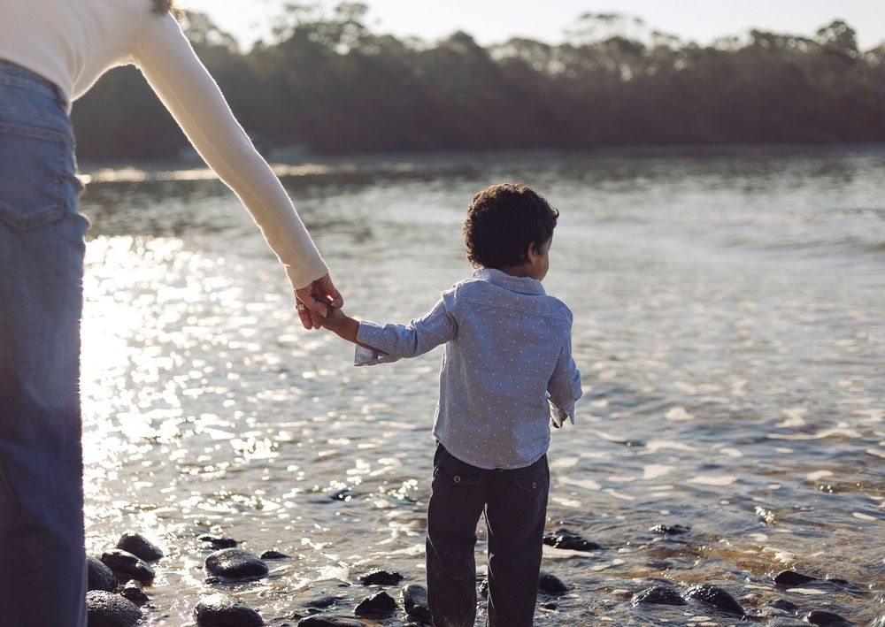 Small child holding an adult's hand while walking along a rocky beach at sunset with water glimmering in the background.