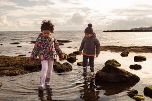 Two figures explore a rocky beach together at sunset while splashing in the shallow coastal waters.