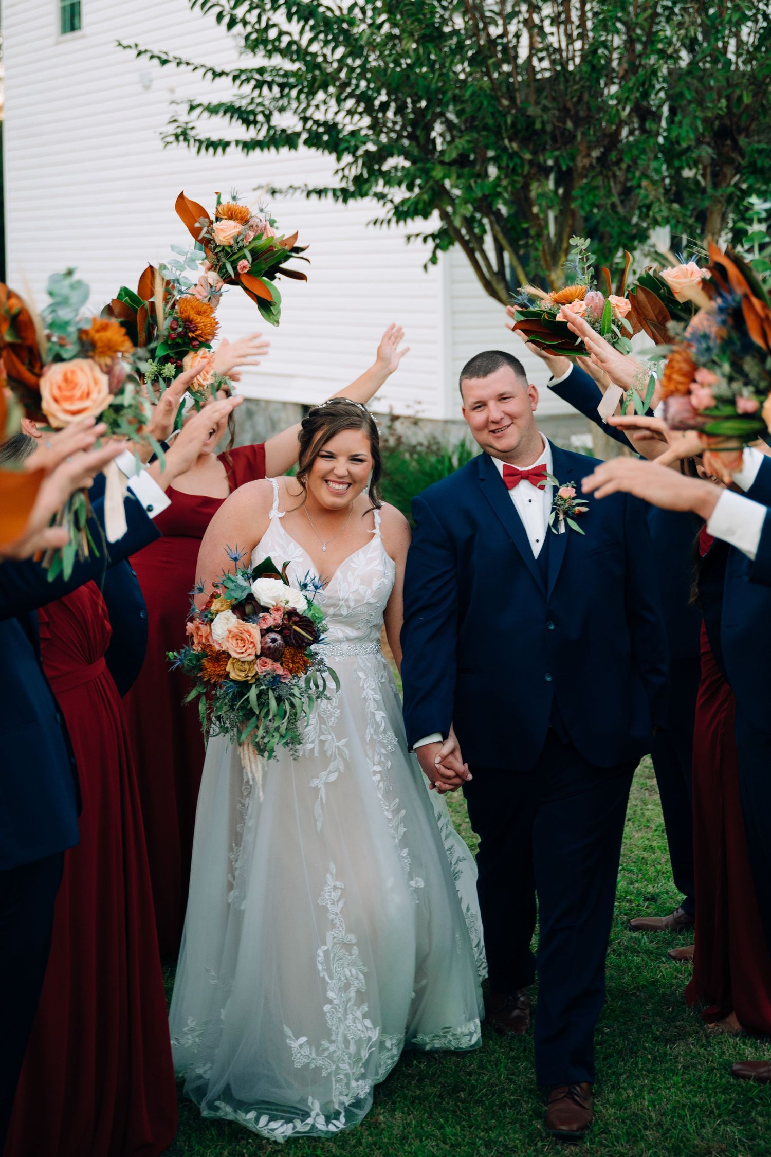 Wedding party celebrates outdoors with raised arms wearing burgundy and formal attire.