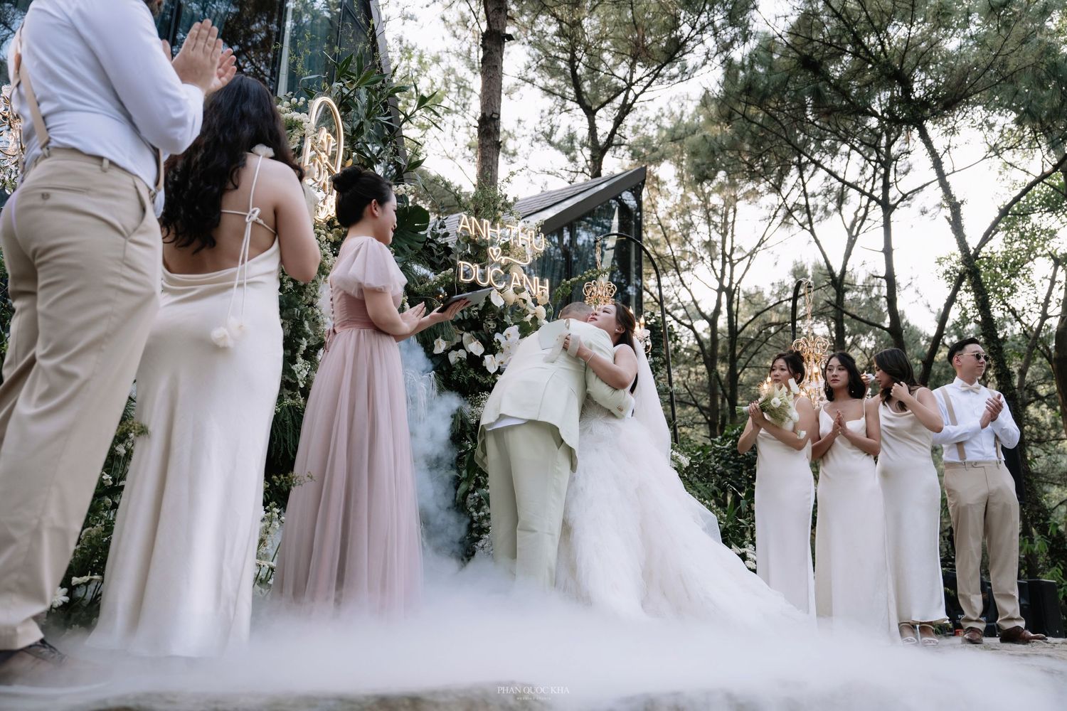 Bride and groom share a first kiss surrounded by bridesmaids and groomsmen in a misty outdoor ceremony.