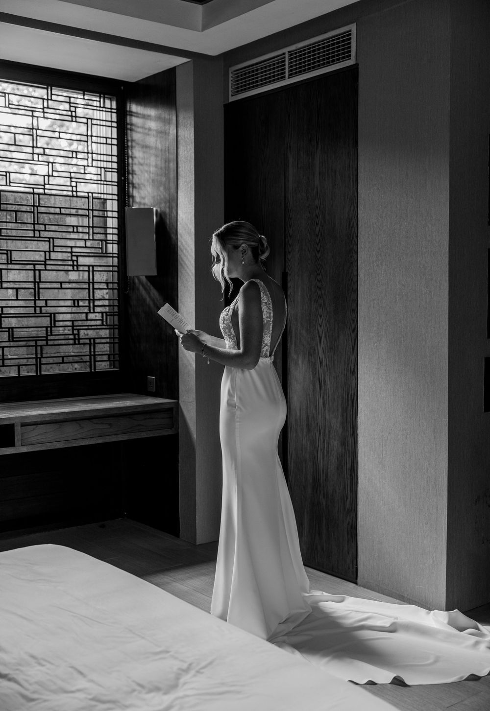 A black and white photo of someone in an elegant white wedding dress standing near a window in dramatic lighting.