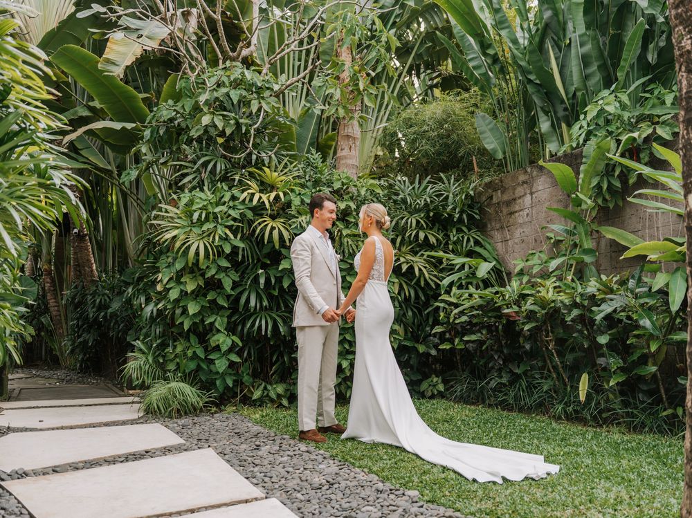 A couple in white attire shares an intimate moment surrounded by lush tropical gardens during their wedding ceremony.