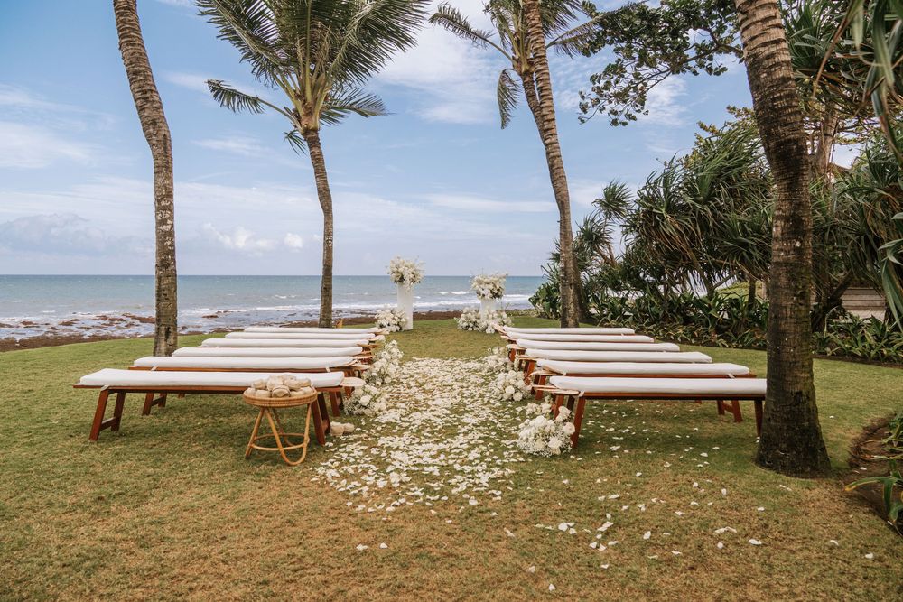 Palm trees sway in the breeze along a tropical beach wedding setup with white benches and scattered flower petals.