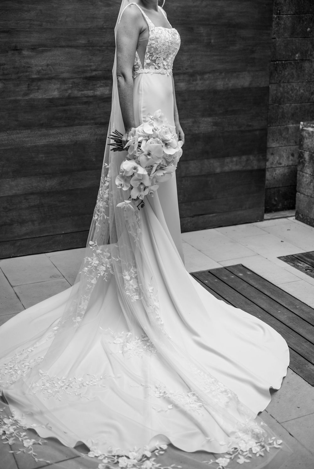 Elegant white wedding dress with lace details and long train displayed against dark wood wall with bride holding orchid bouquet.