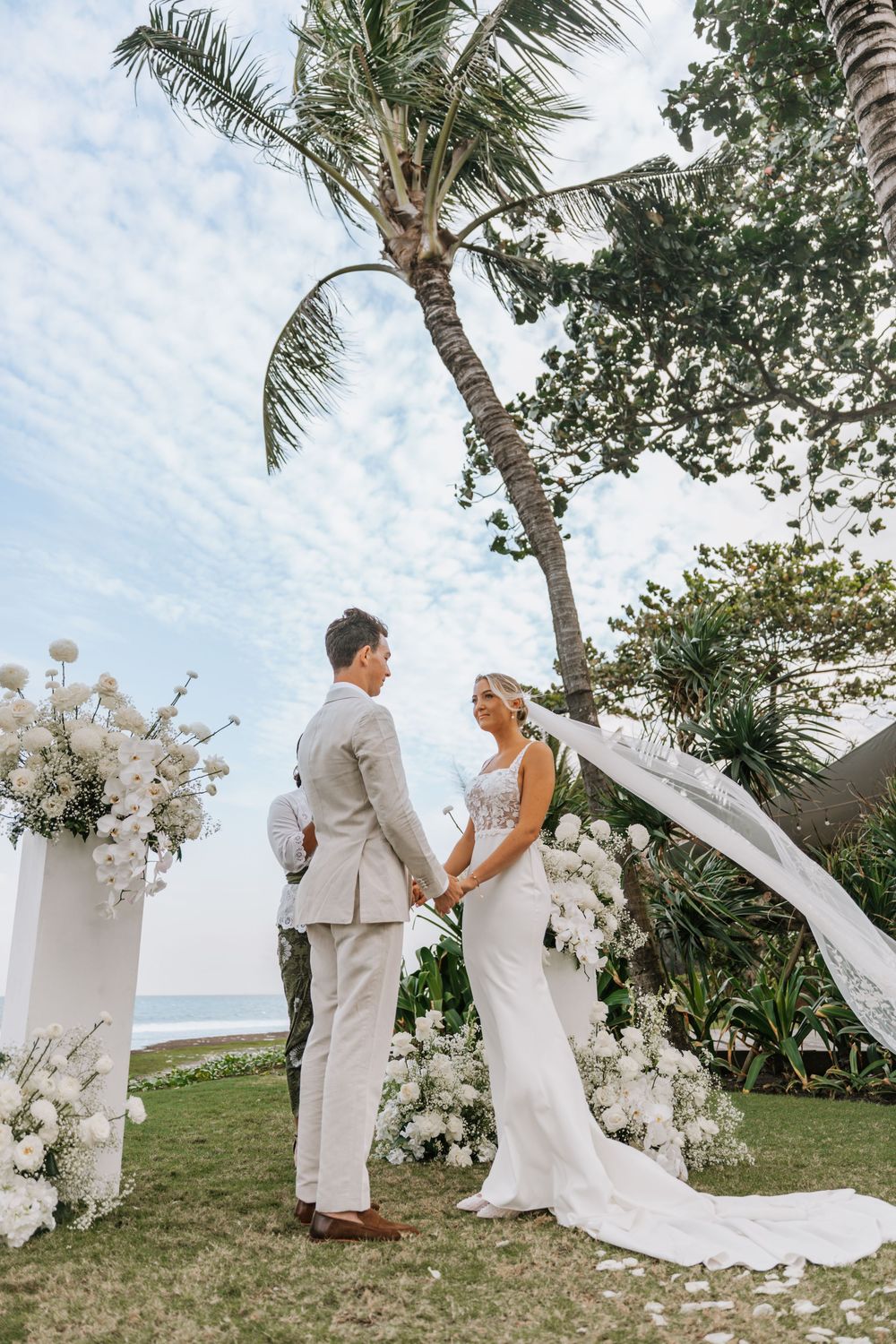 A couple in white and gray attire embrace during their tropical beachside wedding ceremony under palm trees.