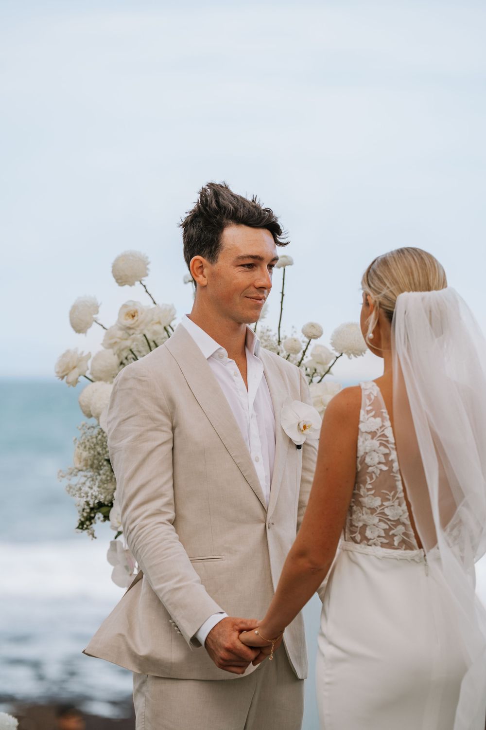 Couple in light beige and white attire share intimate moments during their beachside wedding ceremony with ocean backdrop.
