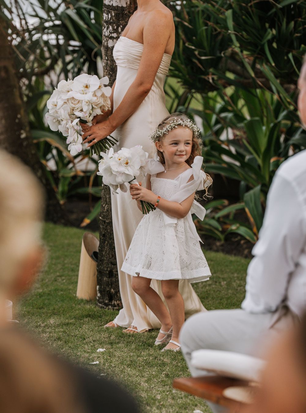 A child in a white dress twirls playfully on a grass lawn during what appears to be an outdoor wedding celebration.