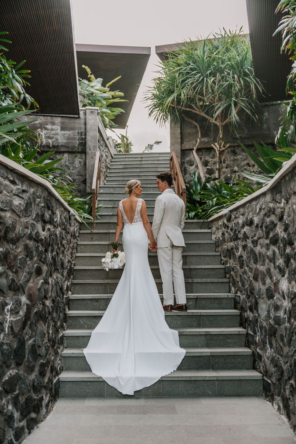 A couple in wedding attire walking up stone stairs surrounded by lush tropical plants and palm trees.