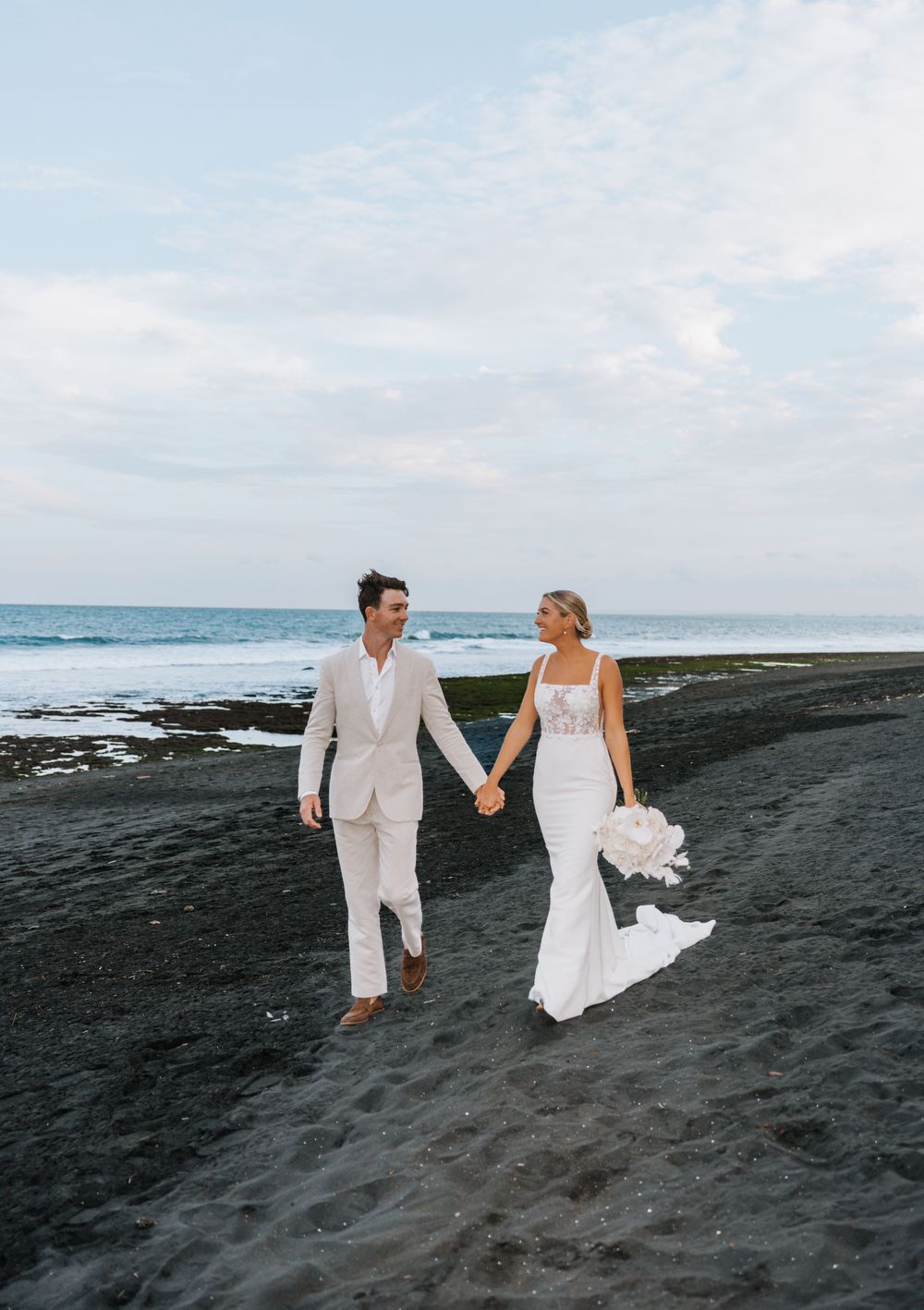 A couple in wedding attire walks hand in hand along a black sand beach with ocean waves and cloudy sky in the background.