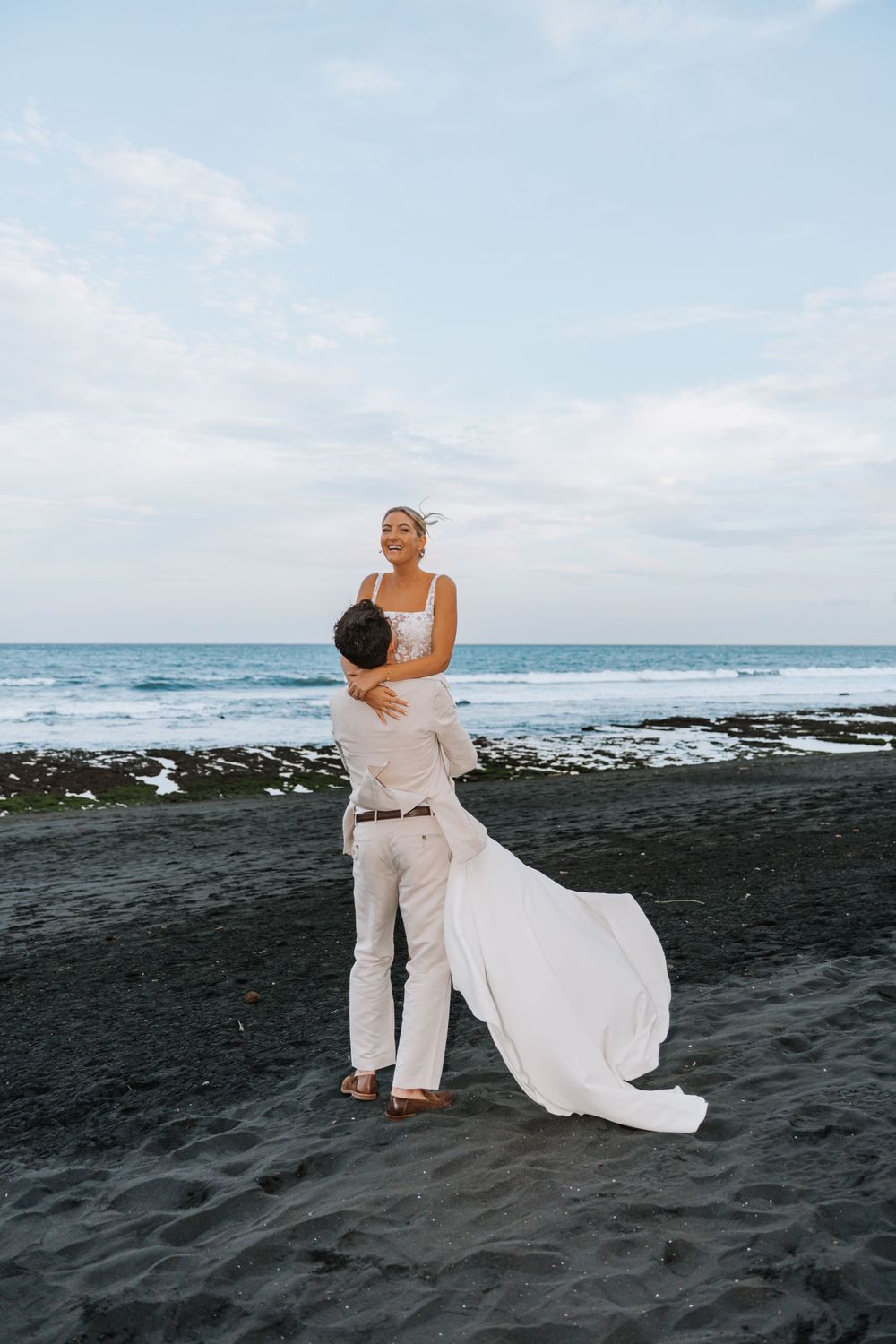 A couple embraces on a black sand beach at sunset with waves and rocky coastline in the background.