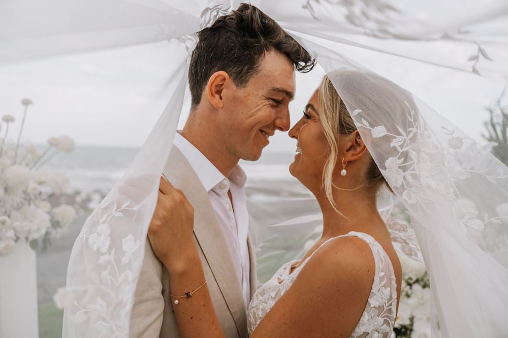 A romantic wedding couple shares an intimate moment under a flowing white veil at an outdoor ceremony.