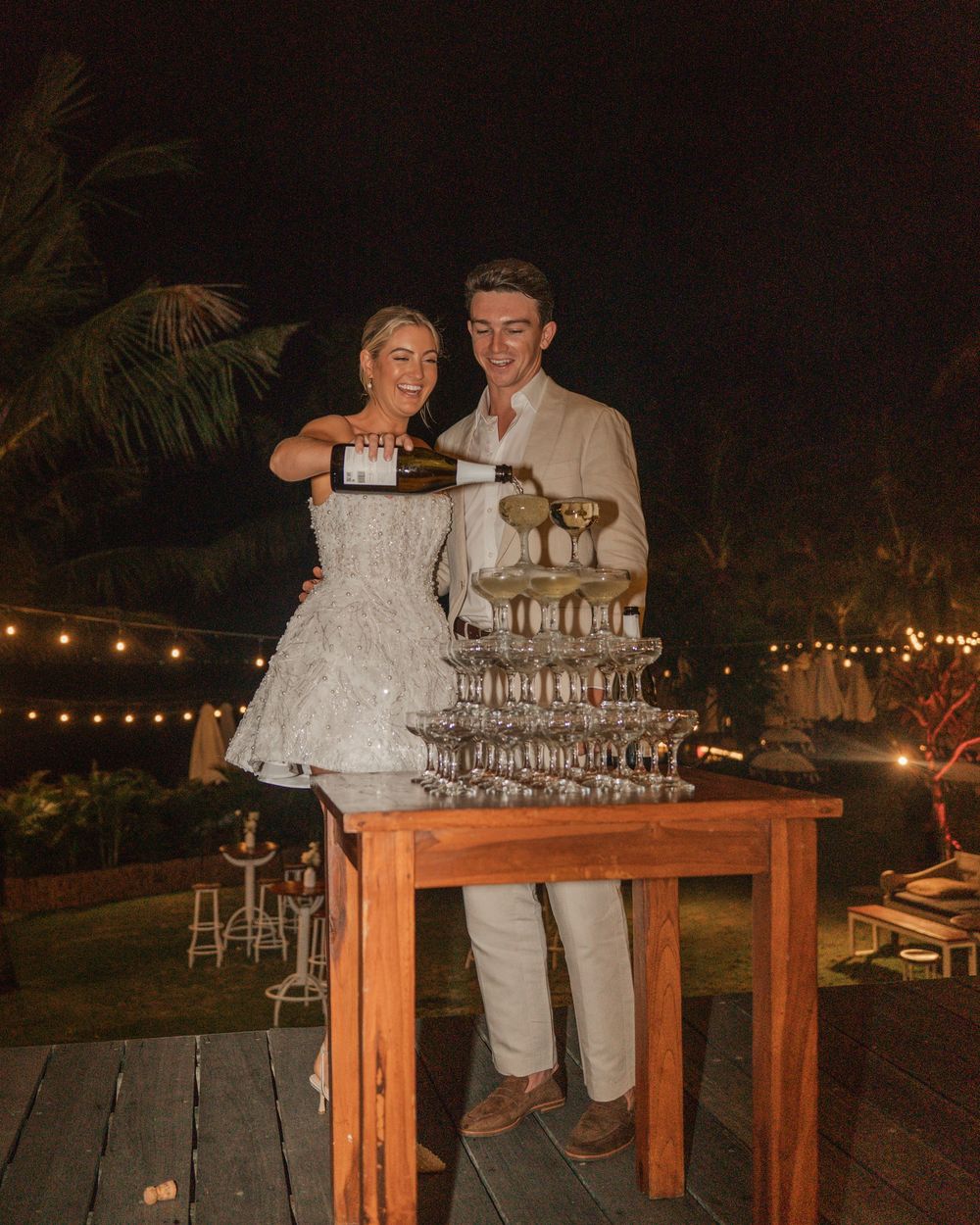 A couple pouring a champagne tower at an outdoor evening wedding reception with string lights in the background.