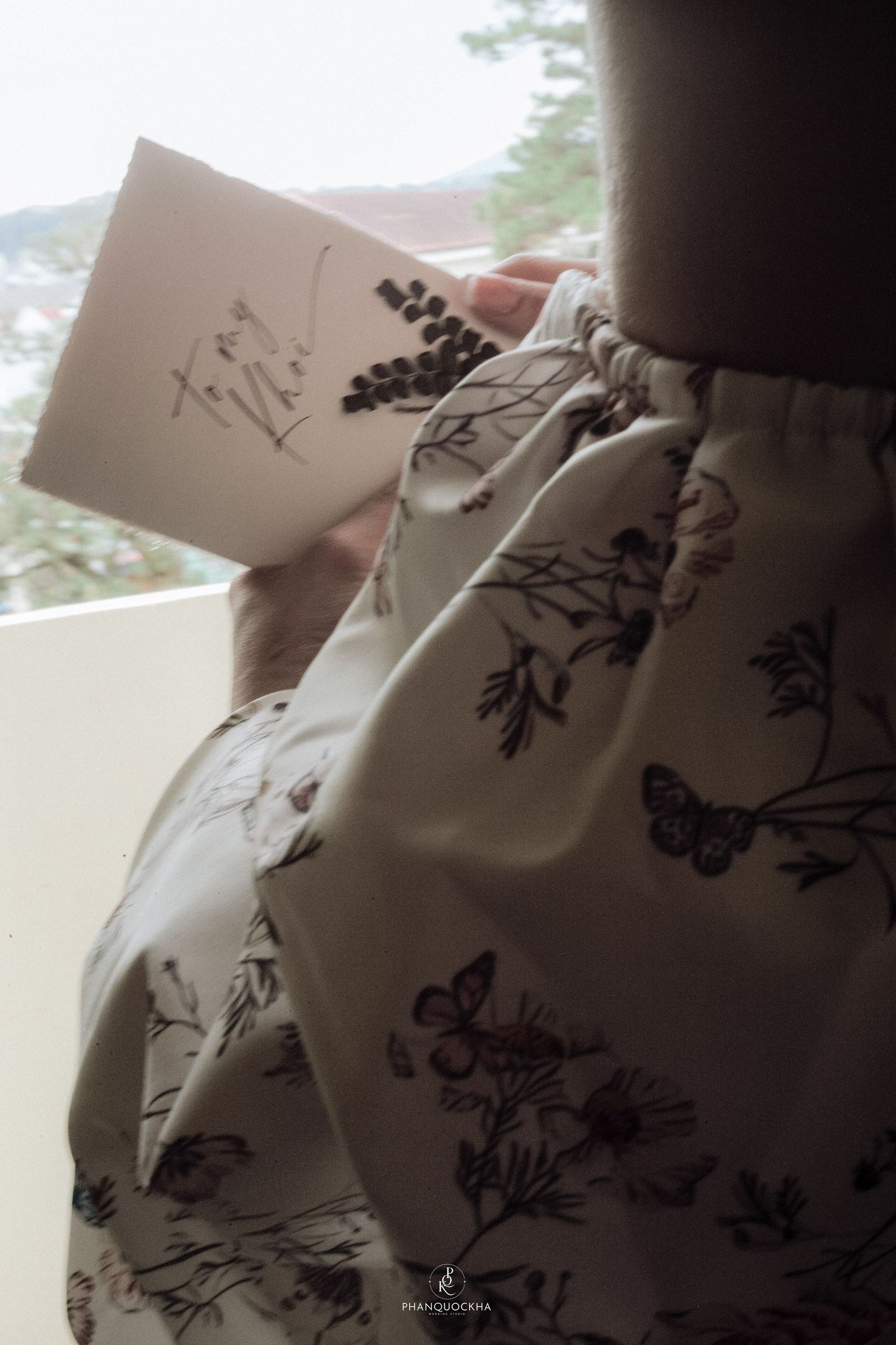 Bride in floral robe reading a wedding card while standing near a bright window.
