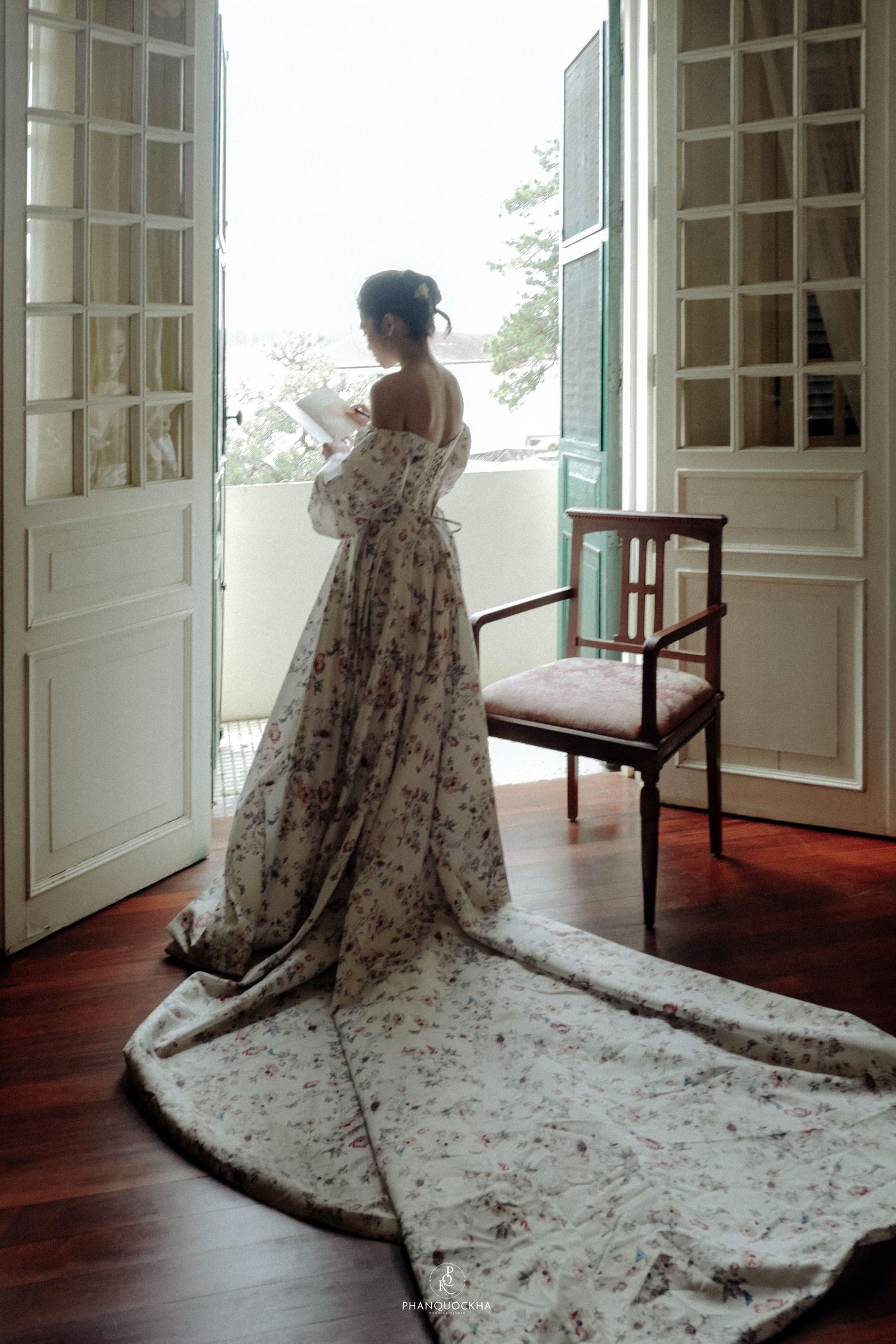 Bride in elegant floral wedding gown with long train standing by open French doors at window.