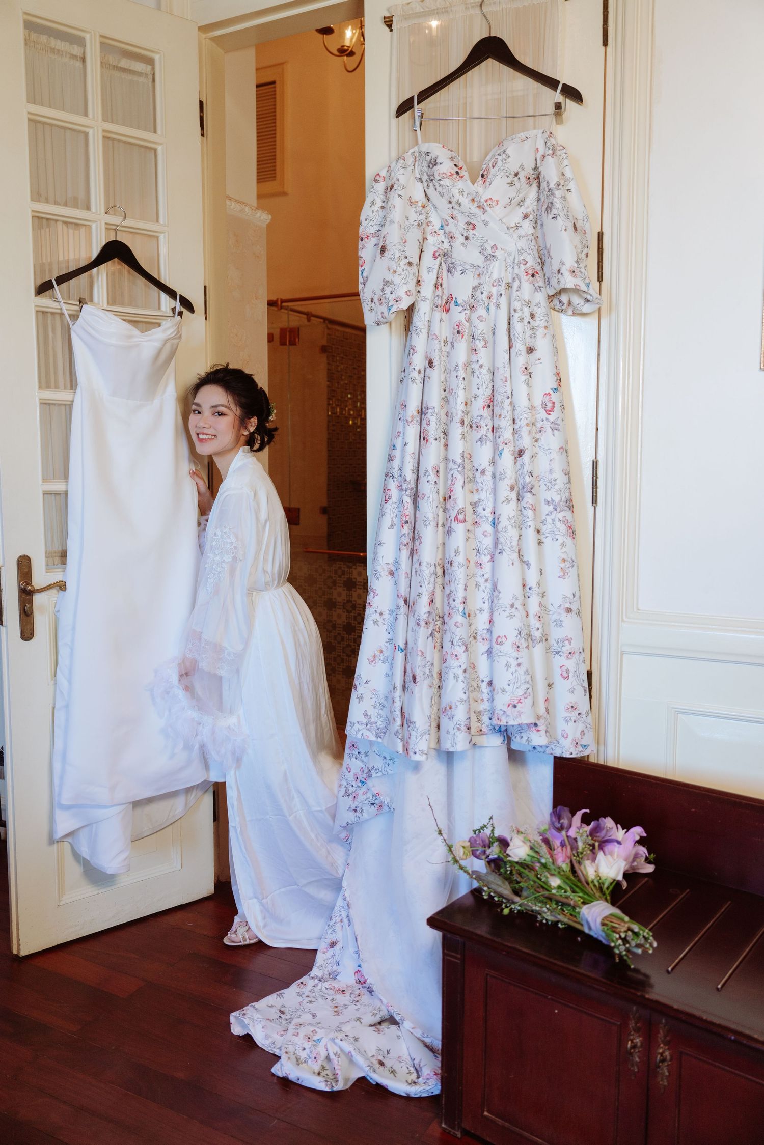 Smiling bride in robe standing between two hanging wedding dresses in a beautifully decorated room.