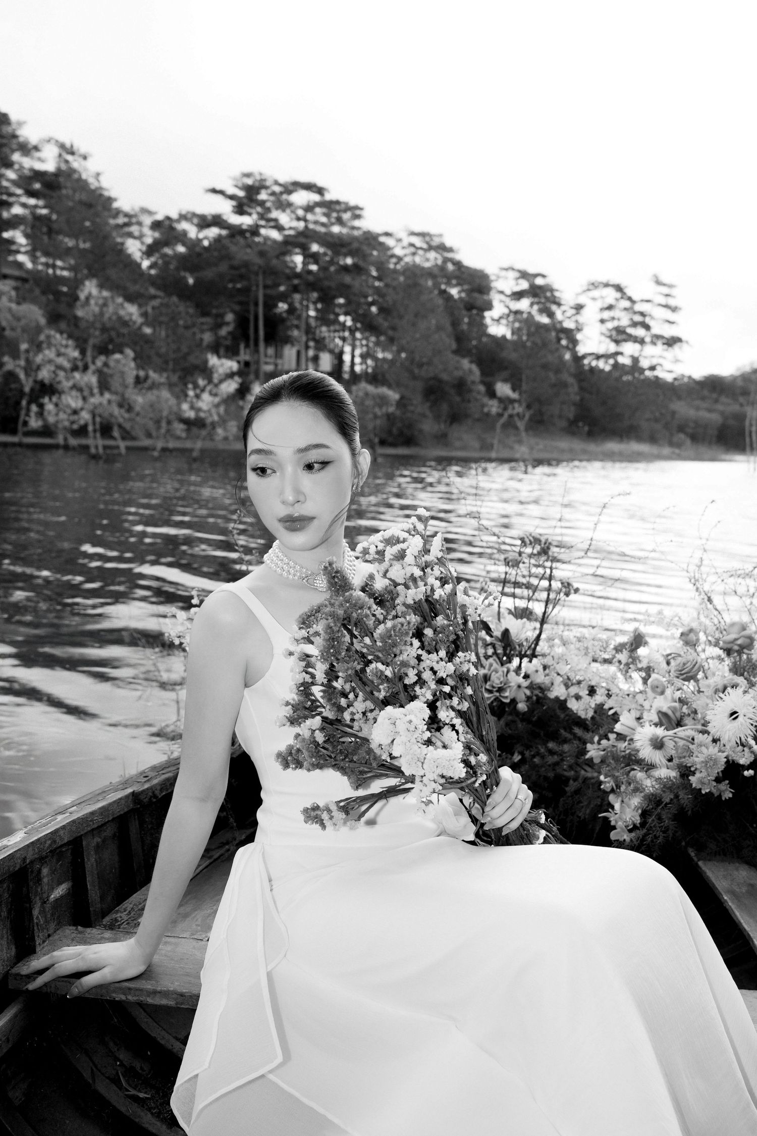 Black and white portrait of elegant bride holding wildflower bouquet in a wooden boat by a lake.