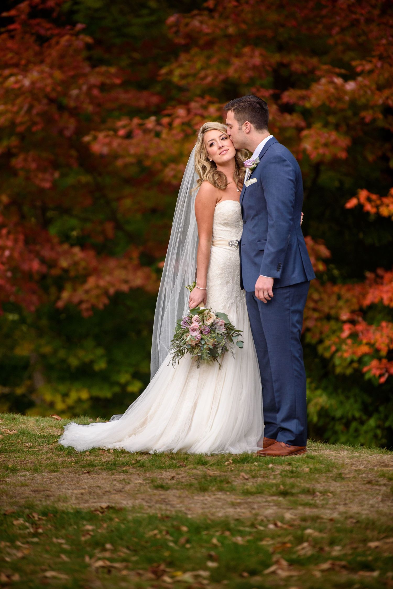 Newlyweds pose against vibrant autumn foliage with bride holding a pink and white bouquet.