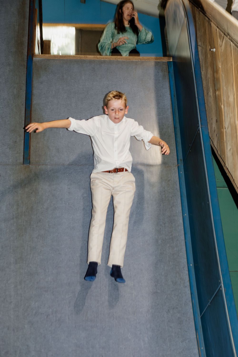 A boy in white clothing slides down a gray ramp in a building with someone standing at the top.