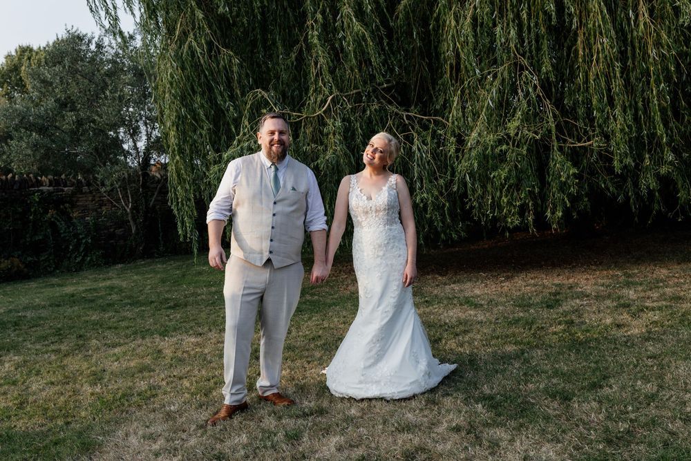 Two people hold hands while standing on grass beneath a drooping willow tree on a sunny summer day.