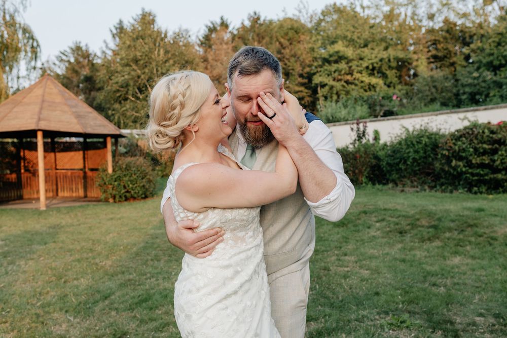 A couple shares a romantic dance on a grassy lawn with rustic wooden buildings in the background at their wedding celebration.