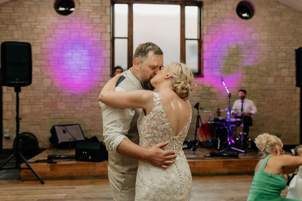A couple shares their first dance together at a rustic brick venue with purple lighting and speakers in the background.