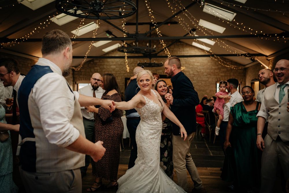 Wedding couple and guests dance together on a rustic wooden dance floor under glowing string lights at an evening reception.