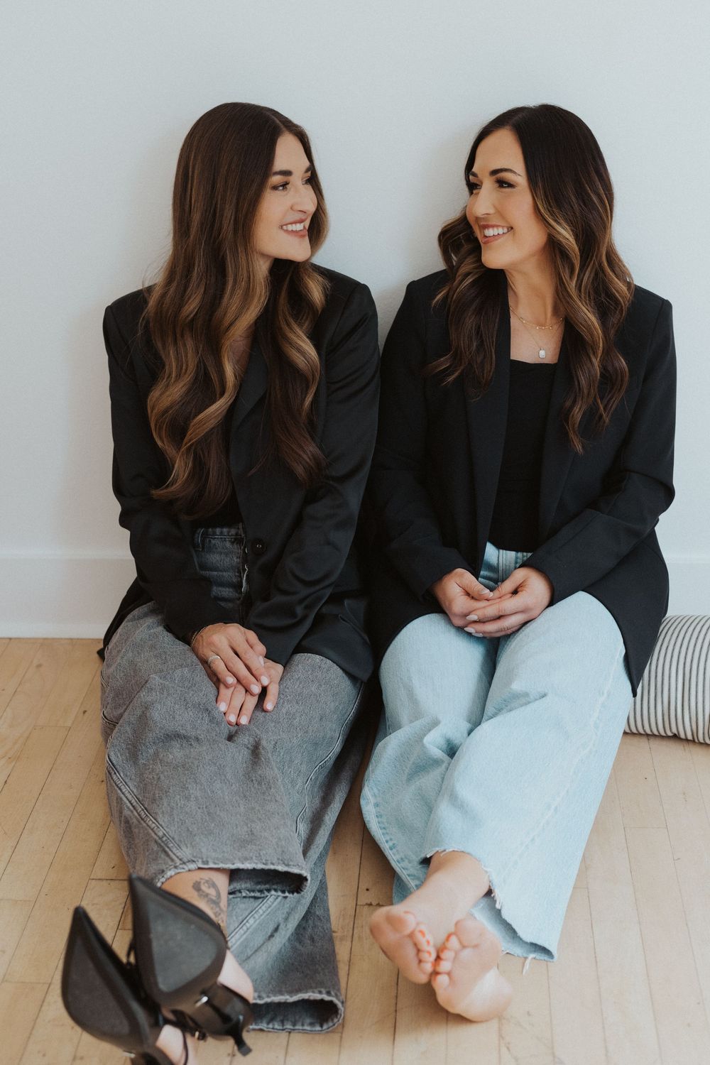 Two people in matching black blazers sitting together on wooden floor for casual photoshoot.