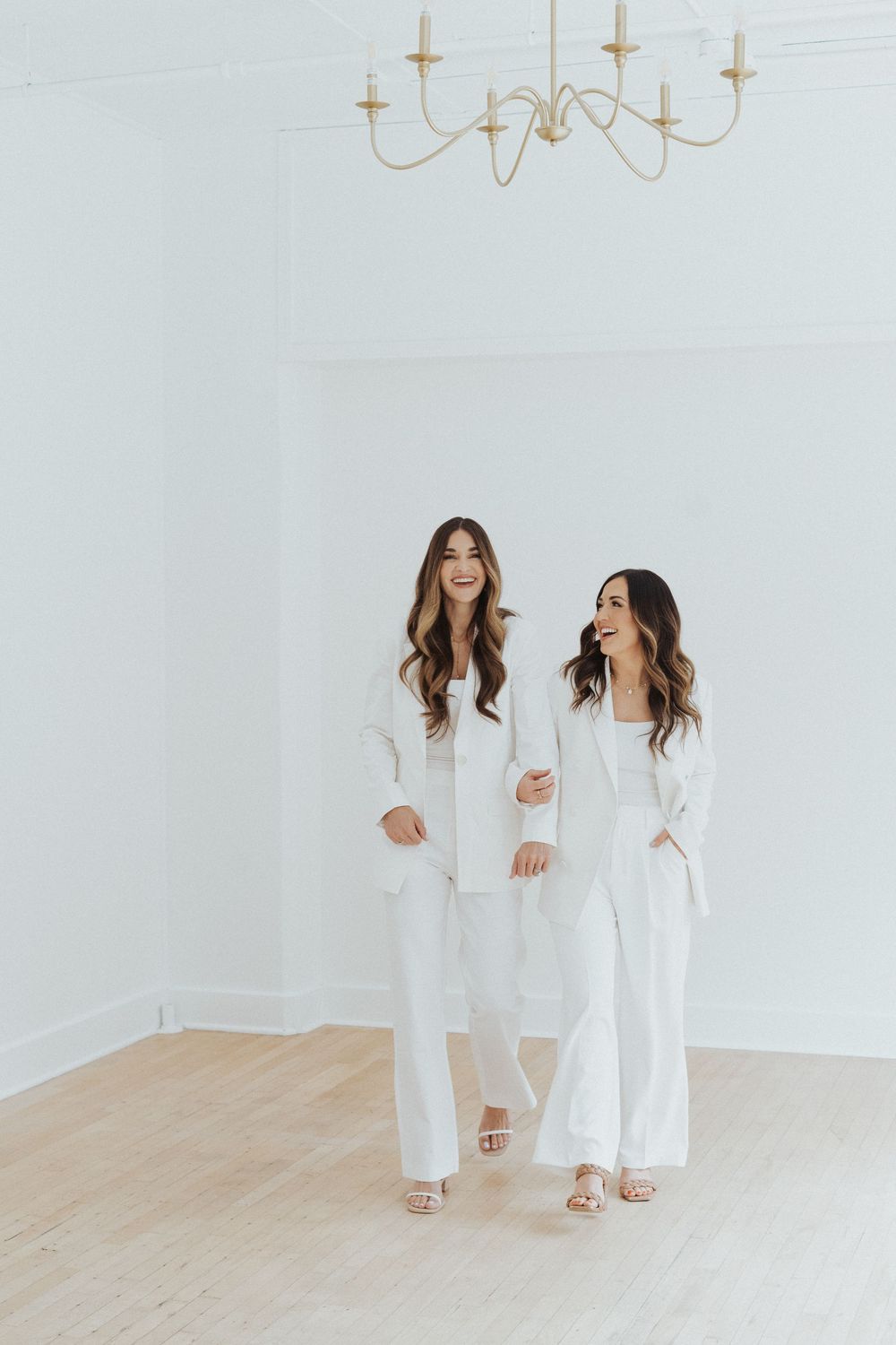 Two people in white outfits standing beneath gold chandelier in bright room.