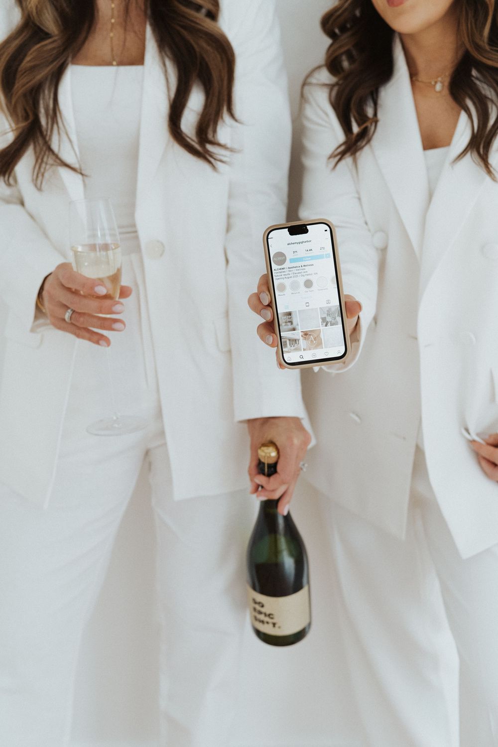 People in white suits holding champagne bottles and phones against white wall.