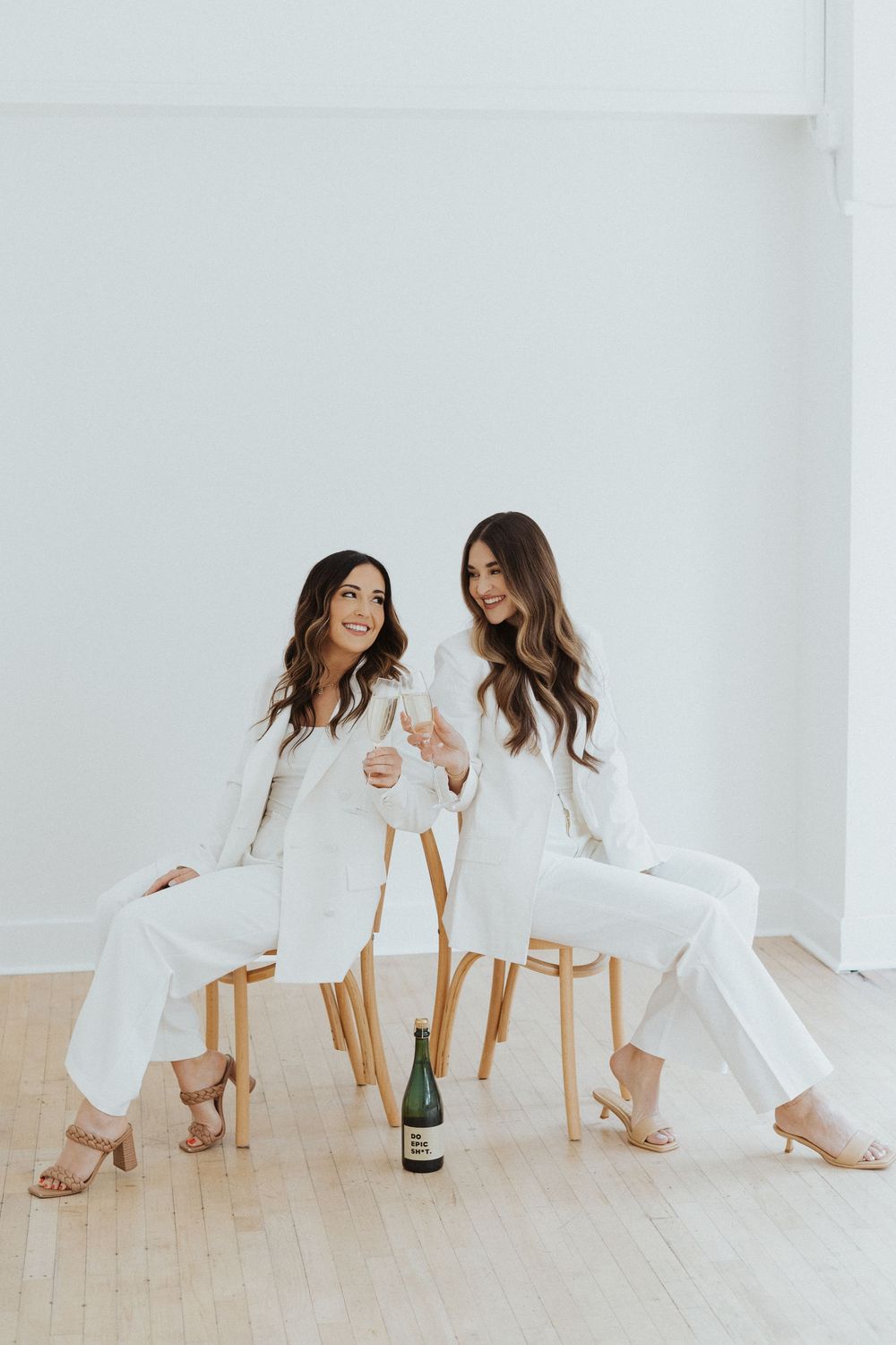 Two people in white outfits sitting on wooden chairs in bright studio setting.