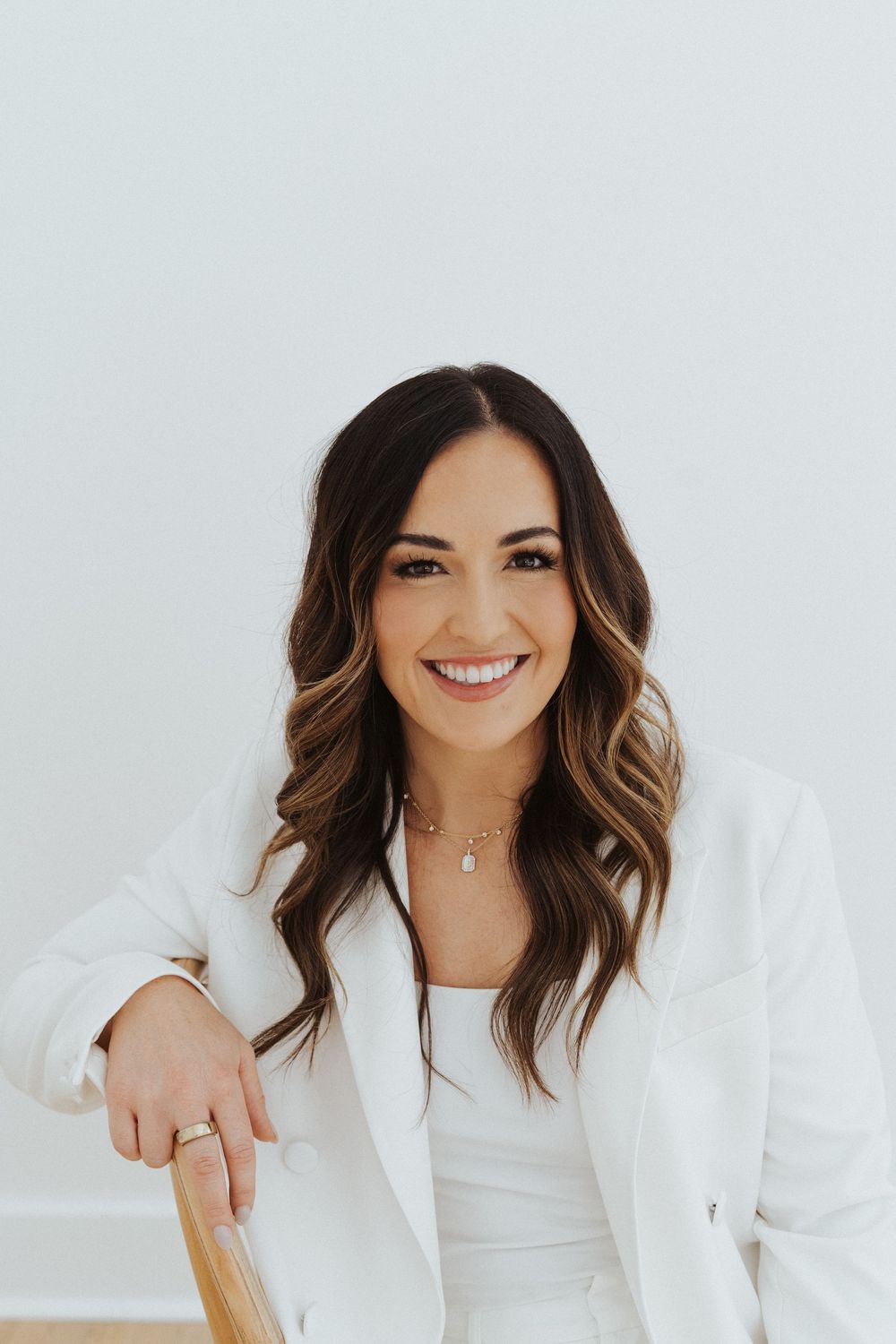 Person in white blazer smiling while leaning on wooden chair in bright studio.