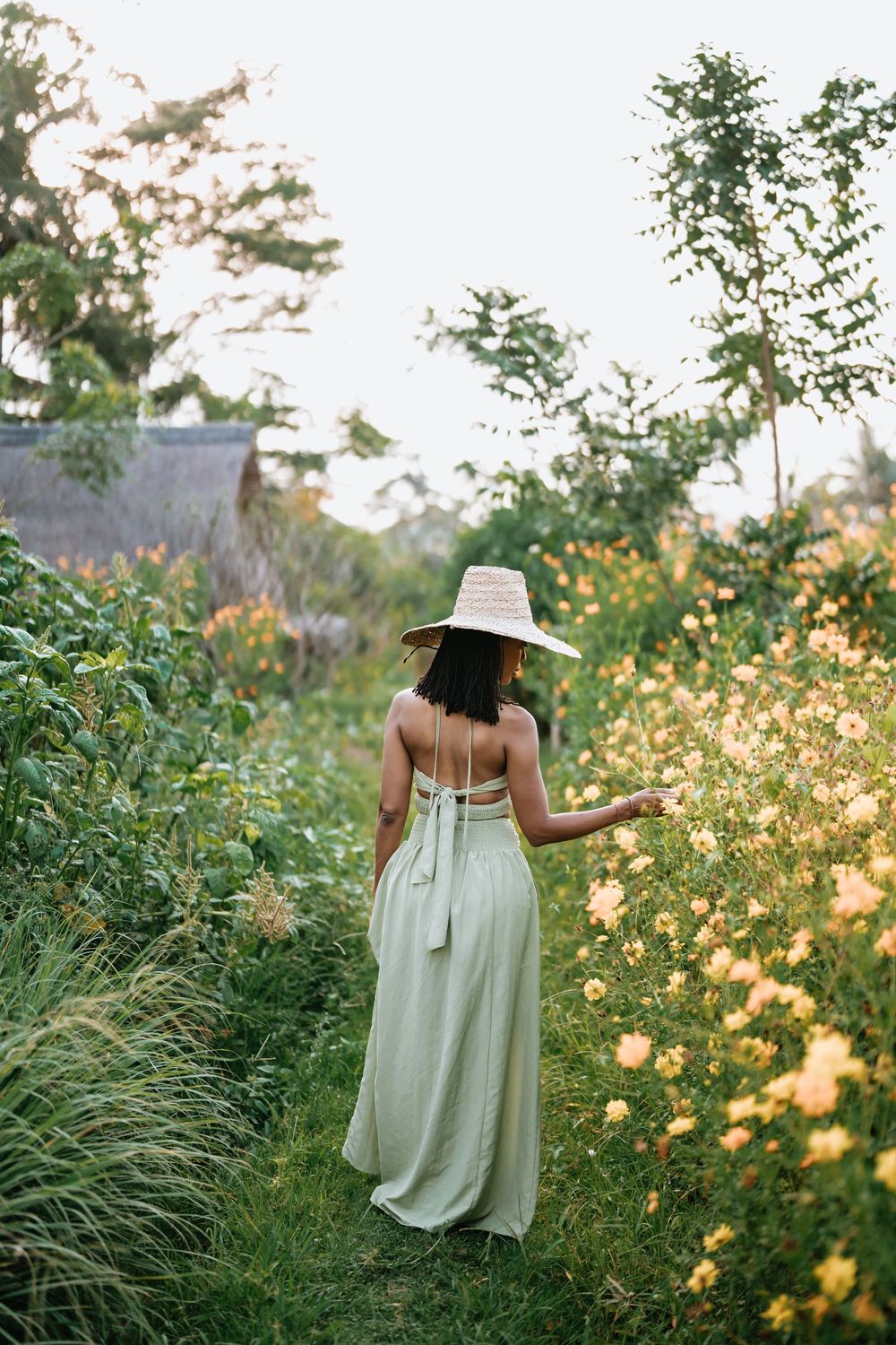 Figure in flowing mint dress and sun hat walking through wildflower field at sunset.