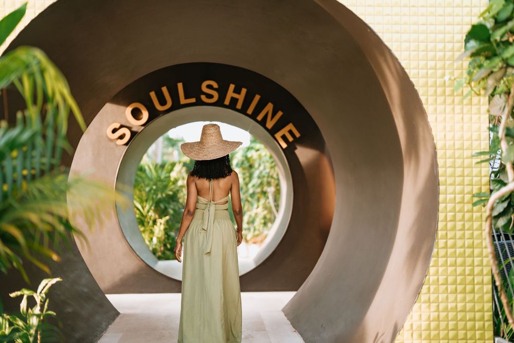 Figure in light dress and hat stands beneath circular Soulshine archway surrounded by tropical plants.