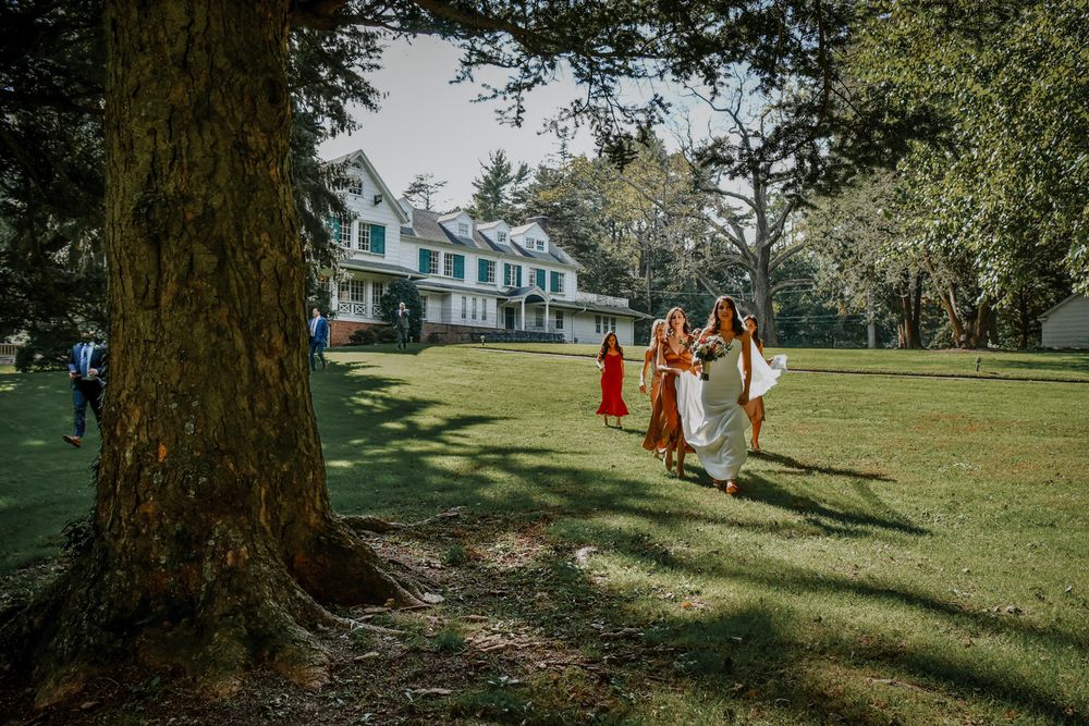A group of people in orange and white dresses walk across a sunlit grassy lawn near a historic mansion.