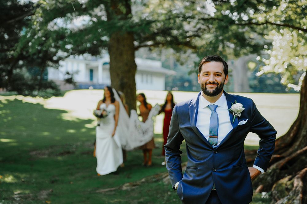 A joyful outdoor wedding scene with a navy suited groom standing in front of trees while a bride follows behind.