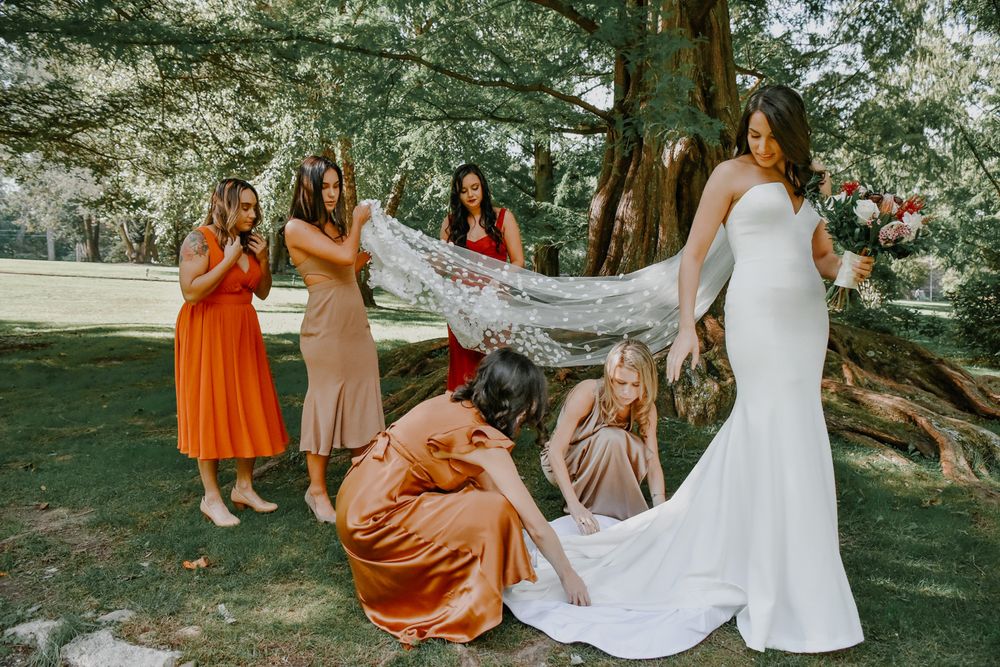 Bridesmaids in orange and blush dresses help adjust white wedding gown on bride outdoors under trees.