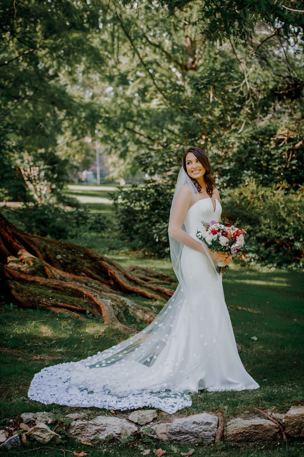 Bride in lace white wedding gown poses gracefully next to rustic fallen tree trunk in lush garden setting.