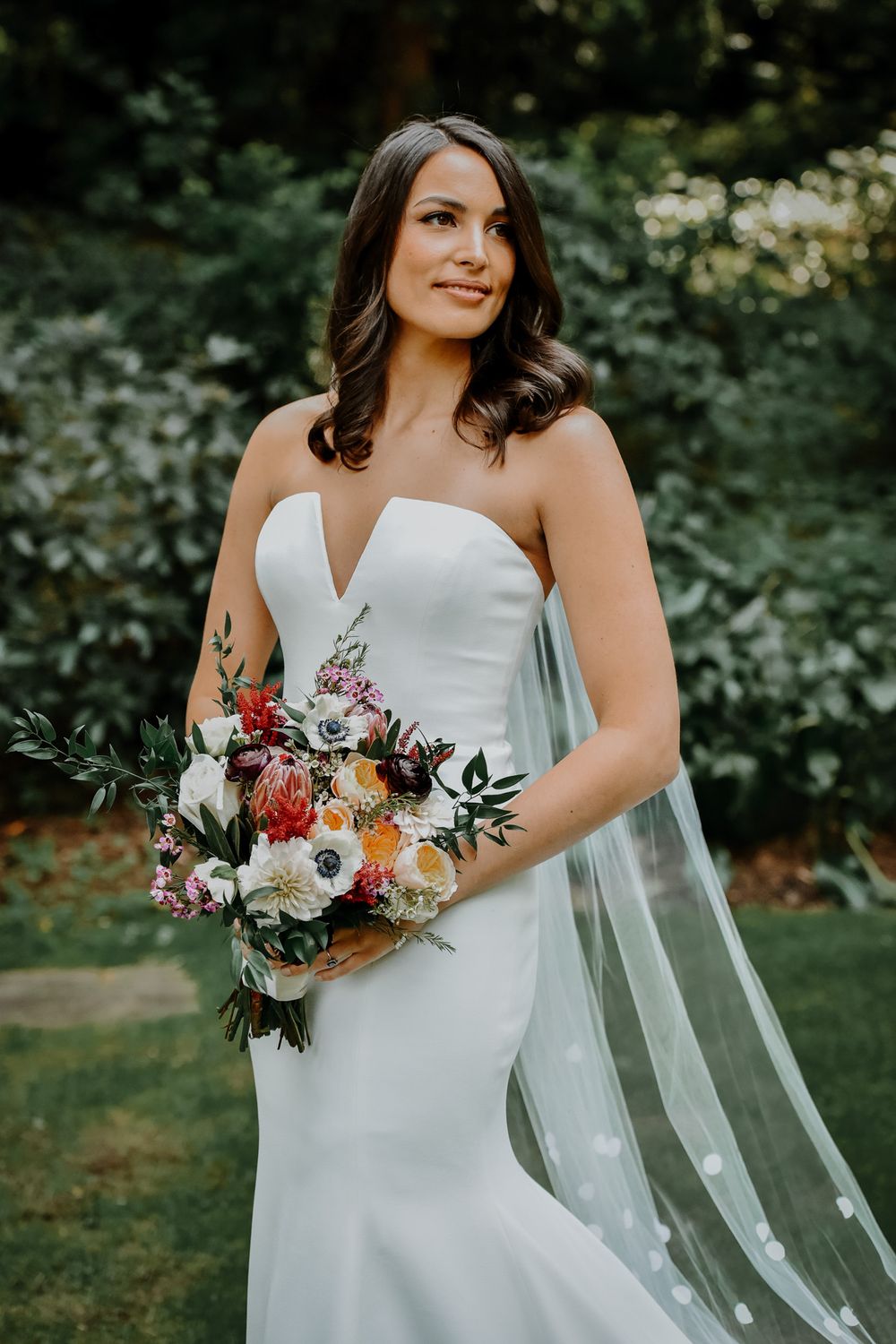 Wedding scene of a bride with flowing white veil holding a colorful bouquet in an outdoor garden setting.