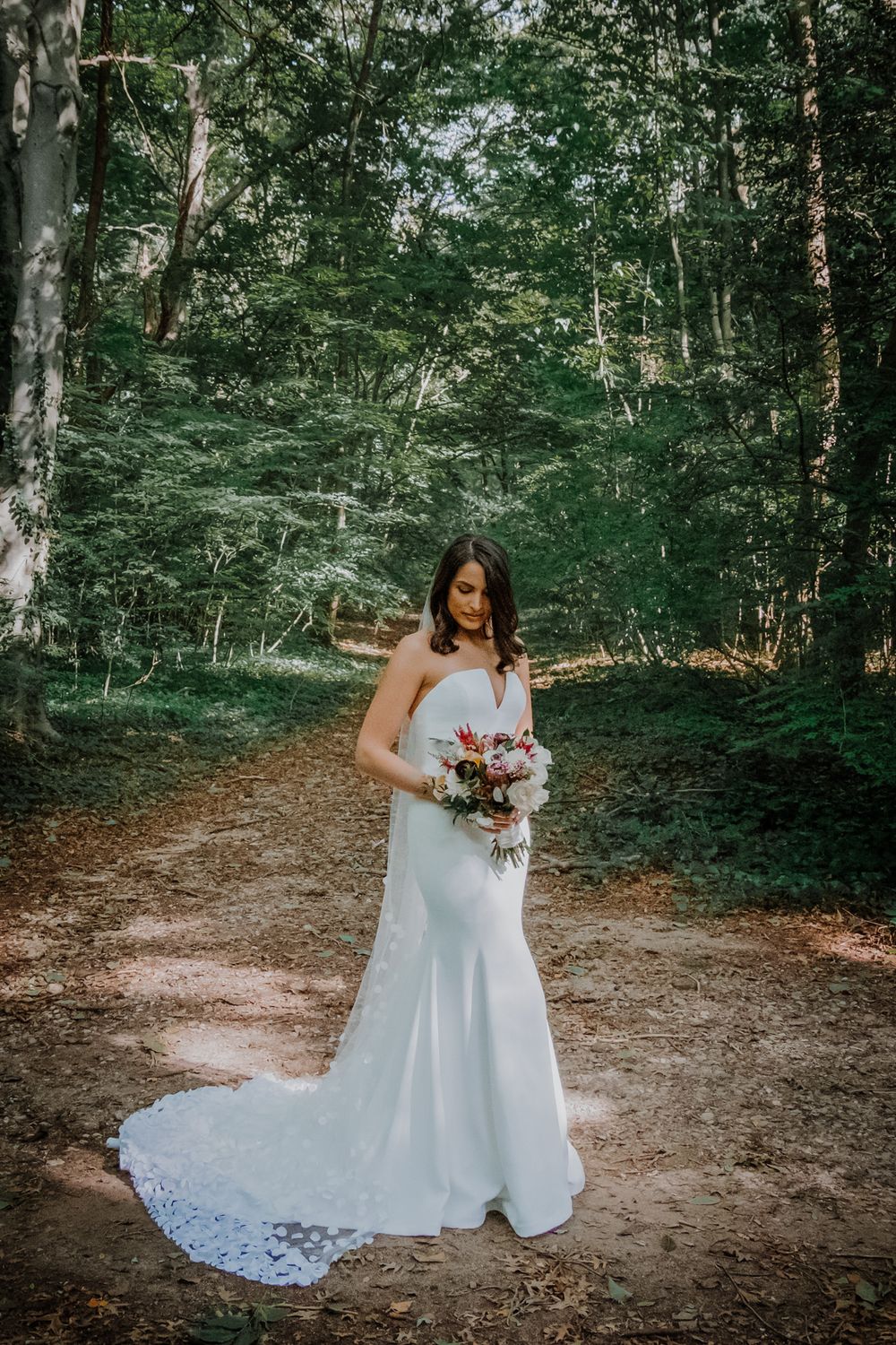 Bride in a white wedding dress poses in a wooded forest setting while holding a colorful bouquet.