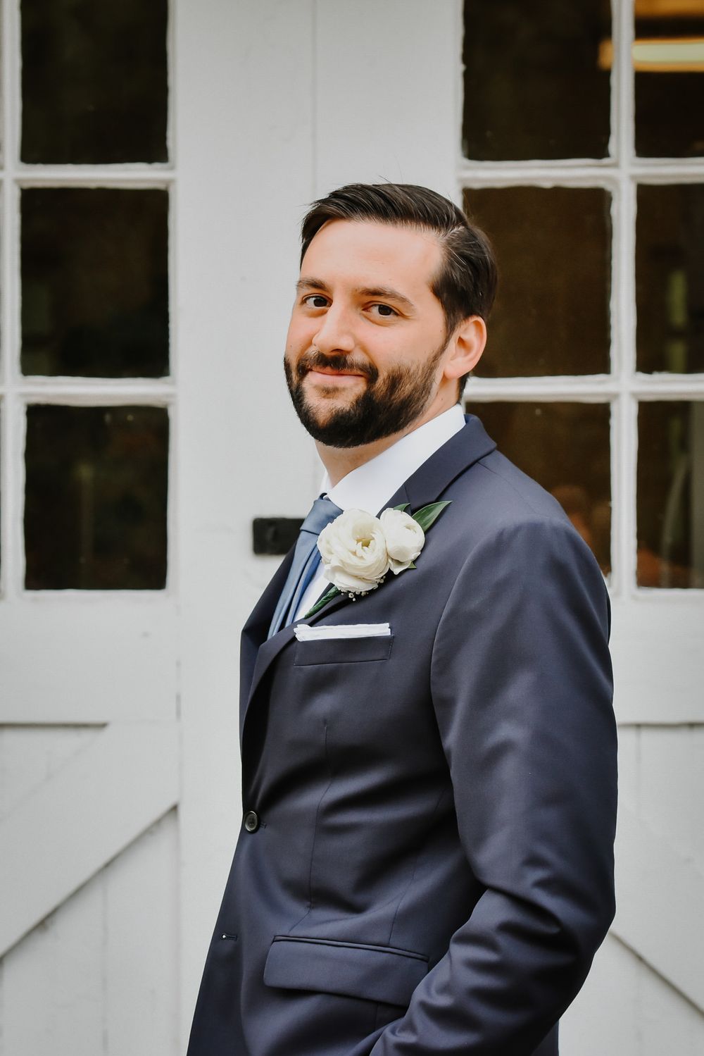 A smiling groom in a navy blue suit with a white boutonniere stands against a white building with glass windows.