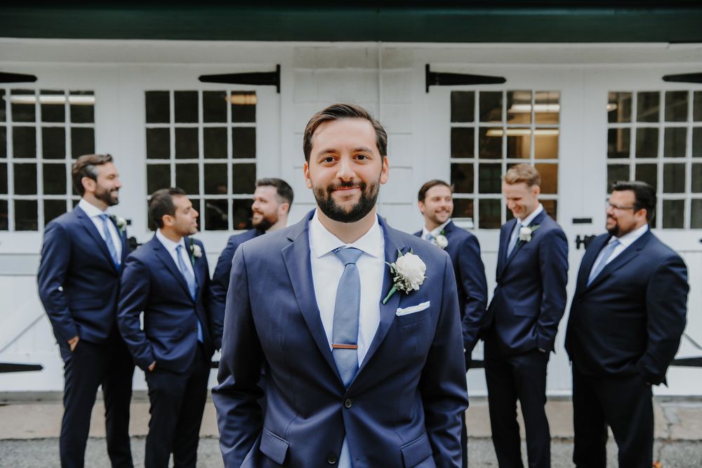 Groomsmen in navy suits stand together outside a building with white windows for wedding party photos.