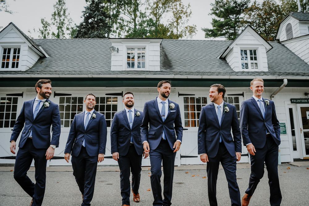 Groomsmen in navy blue suits pose in a line outside a white building with dormers at a wedding celebration.