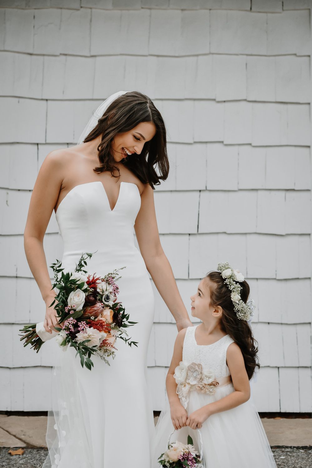 Tender wedding moment showing a bride in white gown and small flower girl in matching dress against a white wall.